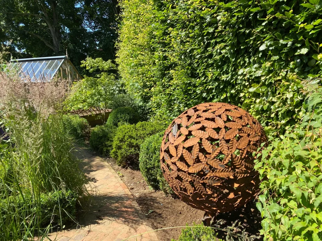 Brass Leaf Sphere Sculpture In Front Of Tall Hedge