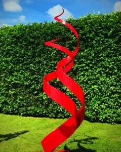 Red Spiral Abstract Sculpture In Front Of Hedge