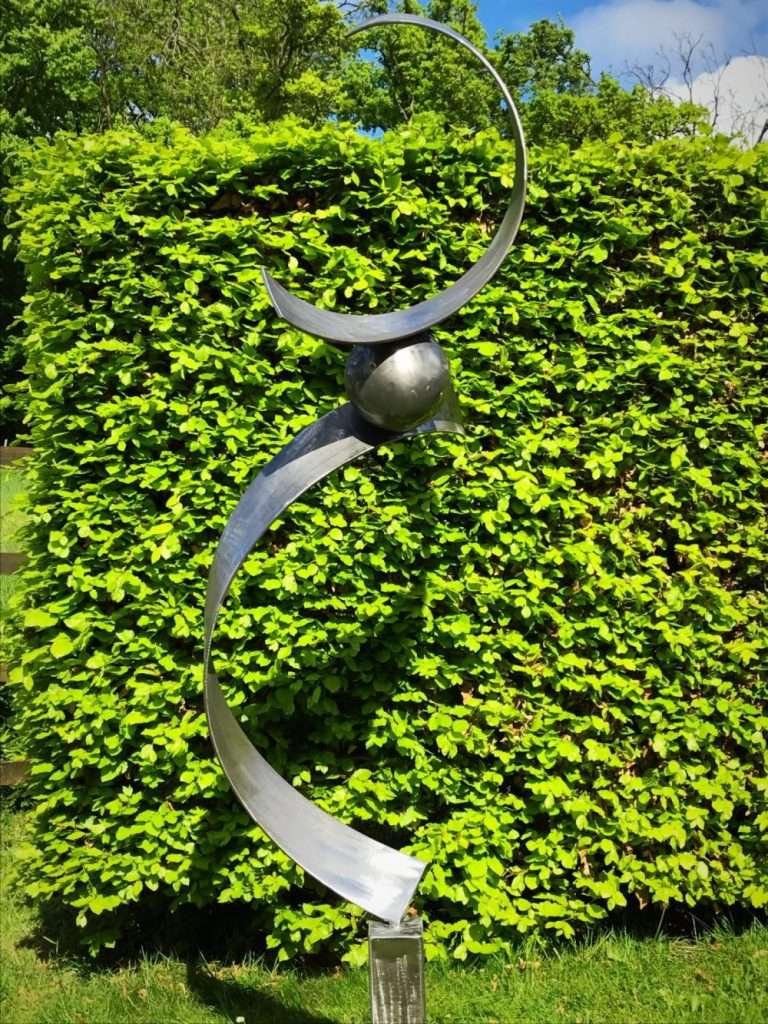large silver abstract sculpture in front of hedge